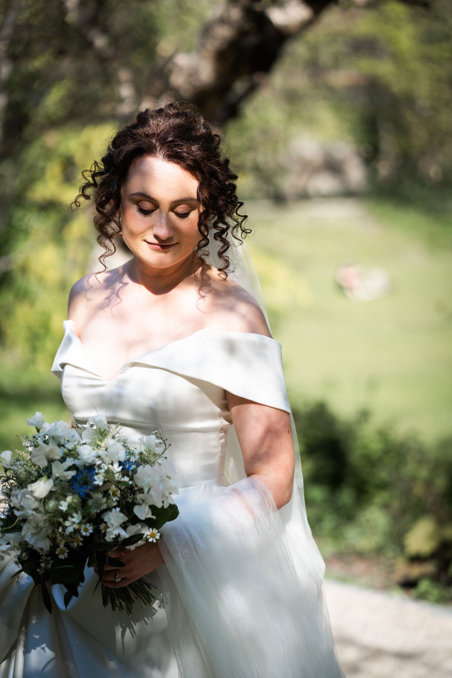 A bride in an off-shoulder white wedding gown holds a blue and white bouquet while looking down on a sunny garden day.