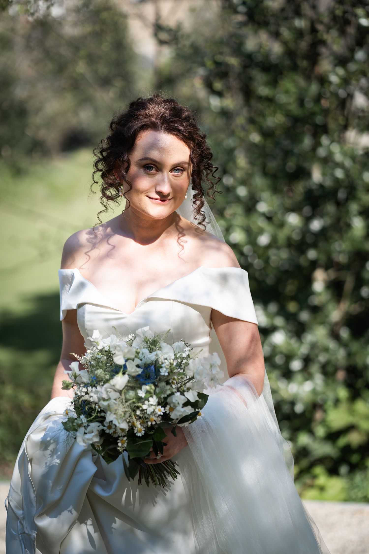 A bride in an off-shoulder white wedding gown holds a delicate bouquet of white and green flowers on a sunny day.