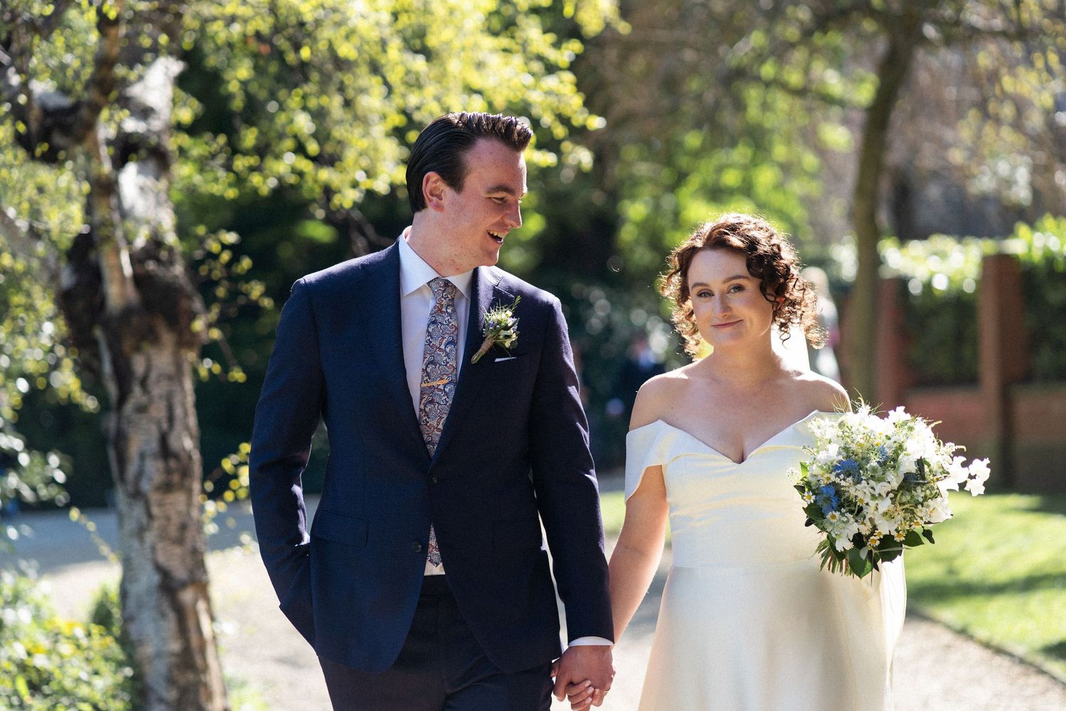 A couple walking together outdoors on their wedding day, with the bride holding a white bouquet.
