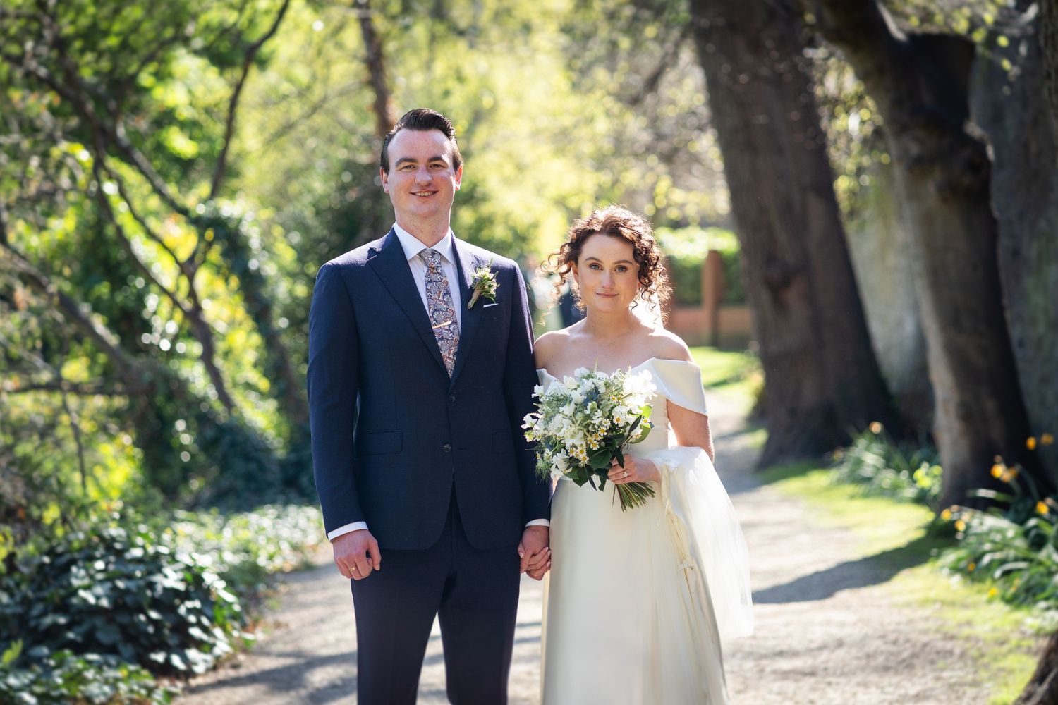 A couple poses together in a scenic garden pathway on their wedding day holding a white floral bouquet.
