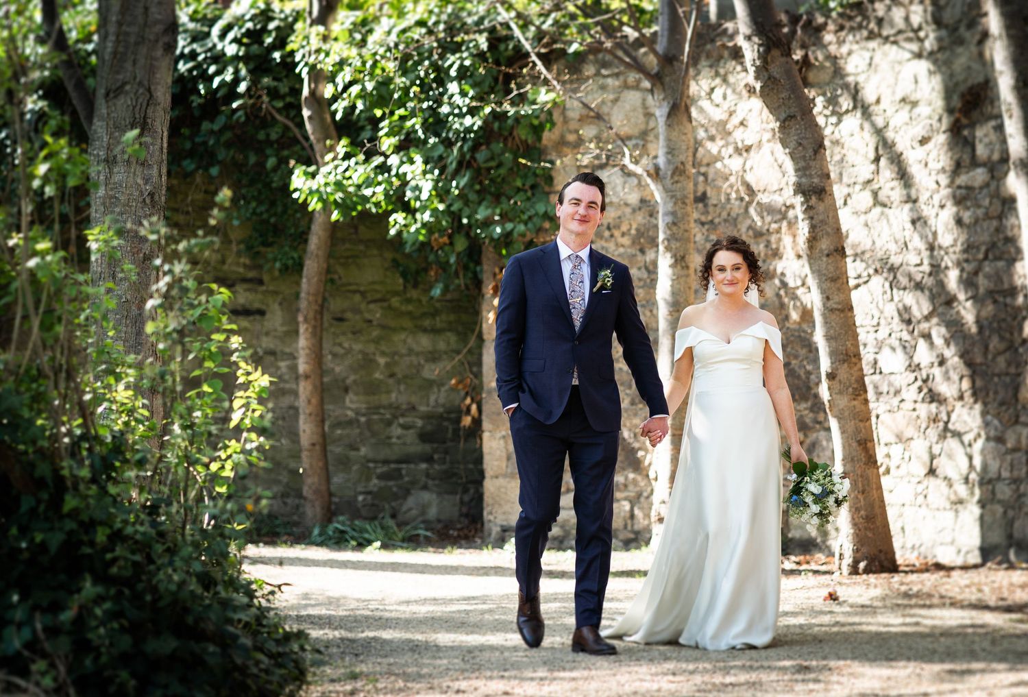 A couple walking hand in hand along a tree-lined path on their wedding day.