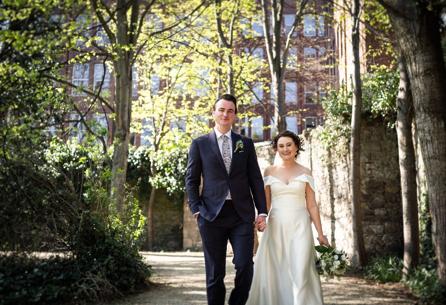 A couple walking down a tree-lined path in formal wedding attire on an autumn day.