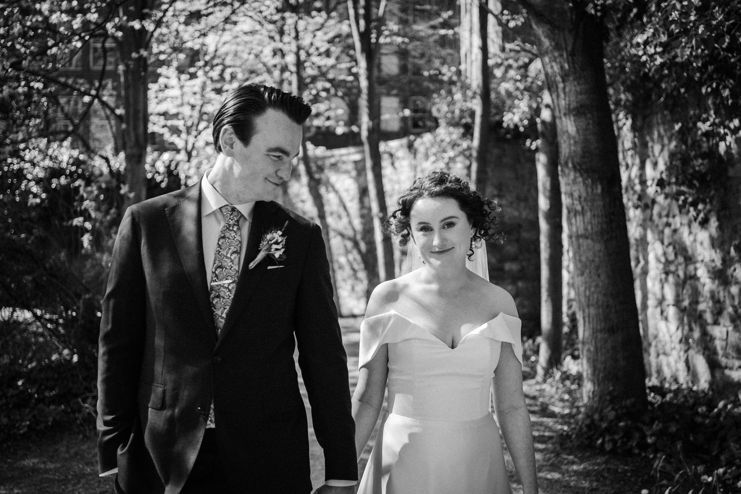 A black and white outdoor wedding photo shows a couple walking together on a wooded path.