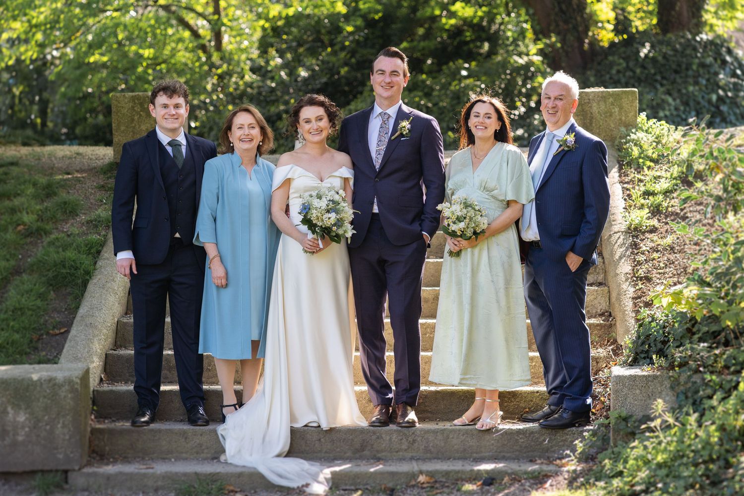 A small wedding party stands together on stone garden steps in a sunlit outdoor setting on a beautiful summer day.