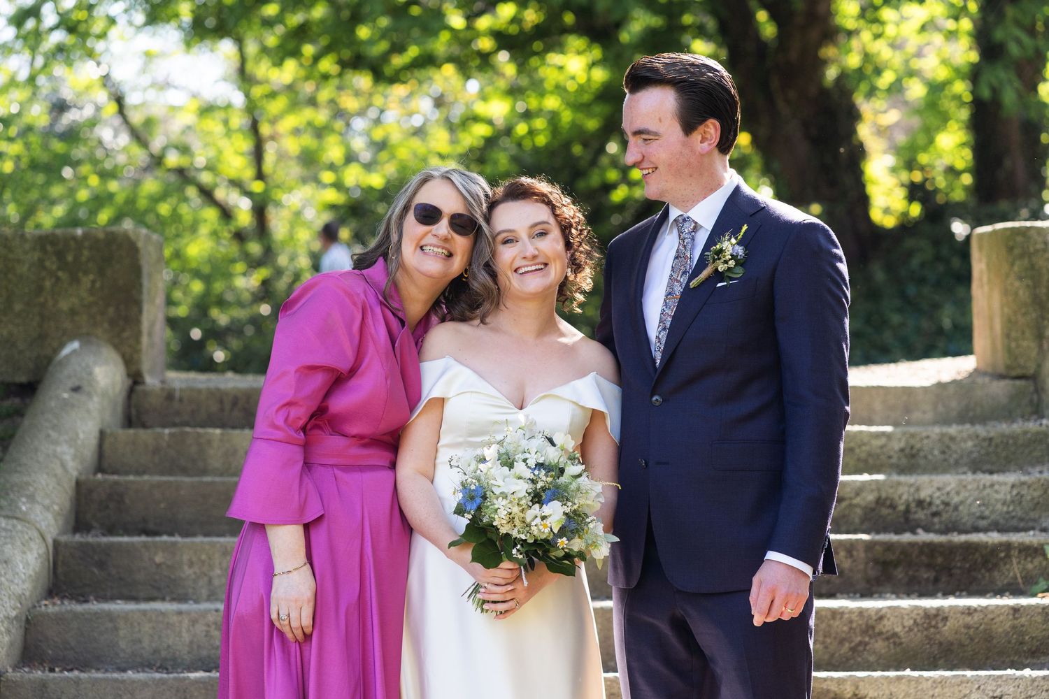 Group photo of three people in wedding attire standing on stone steps outdoors, with bright flowers and greenery in background.