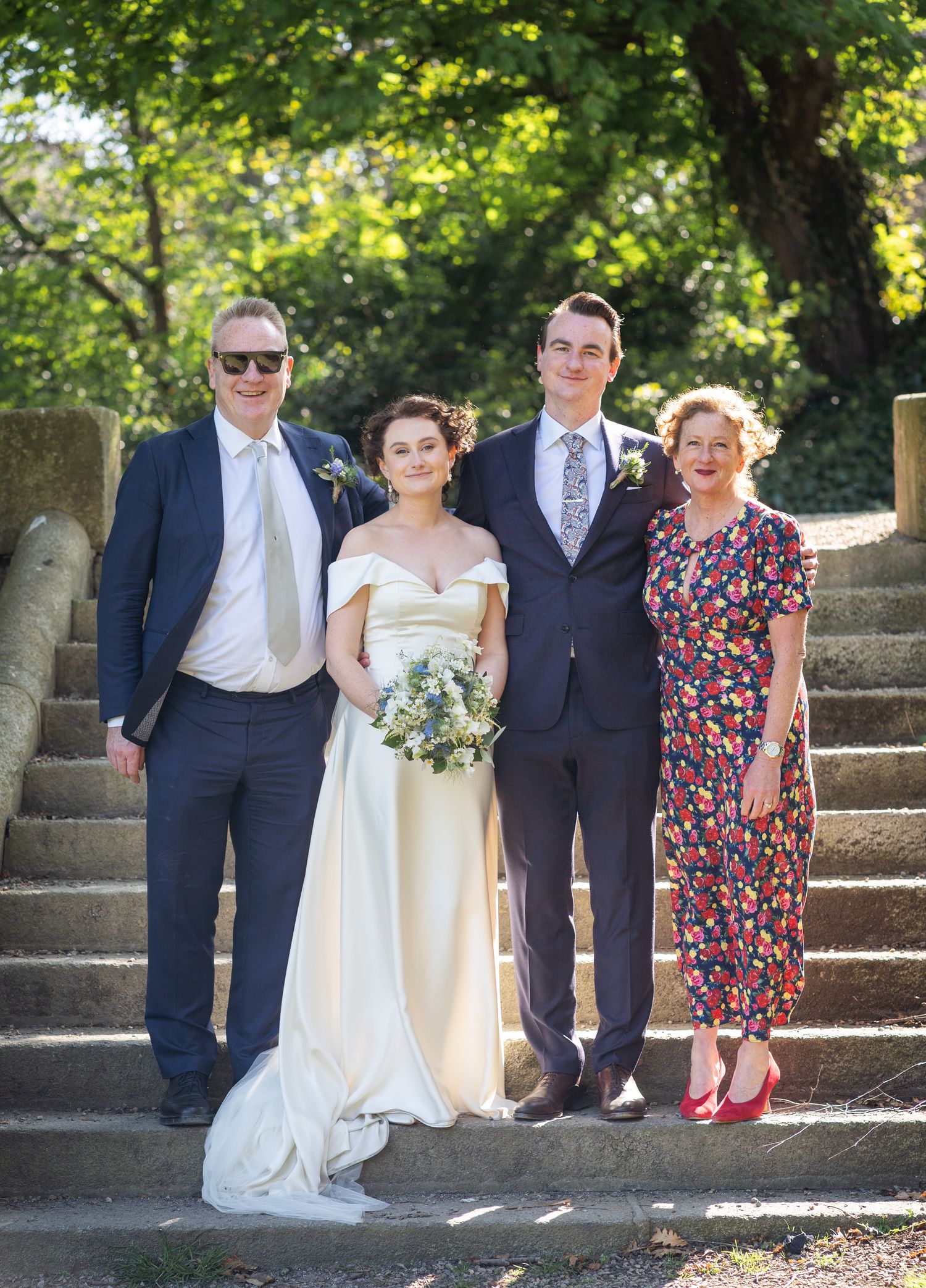 A wedding party of four people poses together outdoors on stone steps surrounded by green foliage on a sunny day.