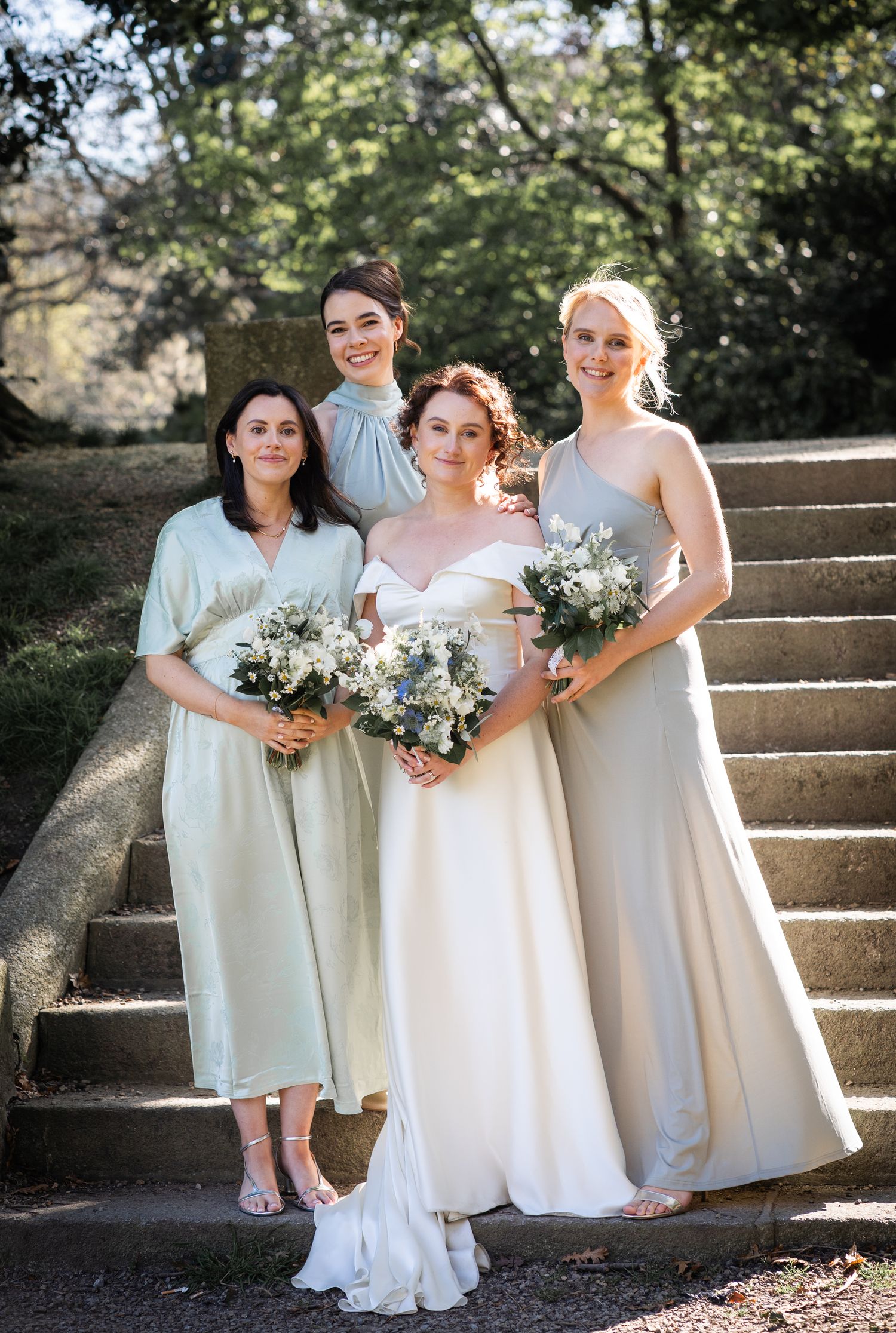 Wedding party members in light-colored dresses pose while holding bouquets on stone steps outdoors.