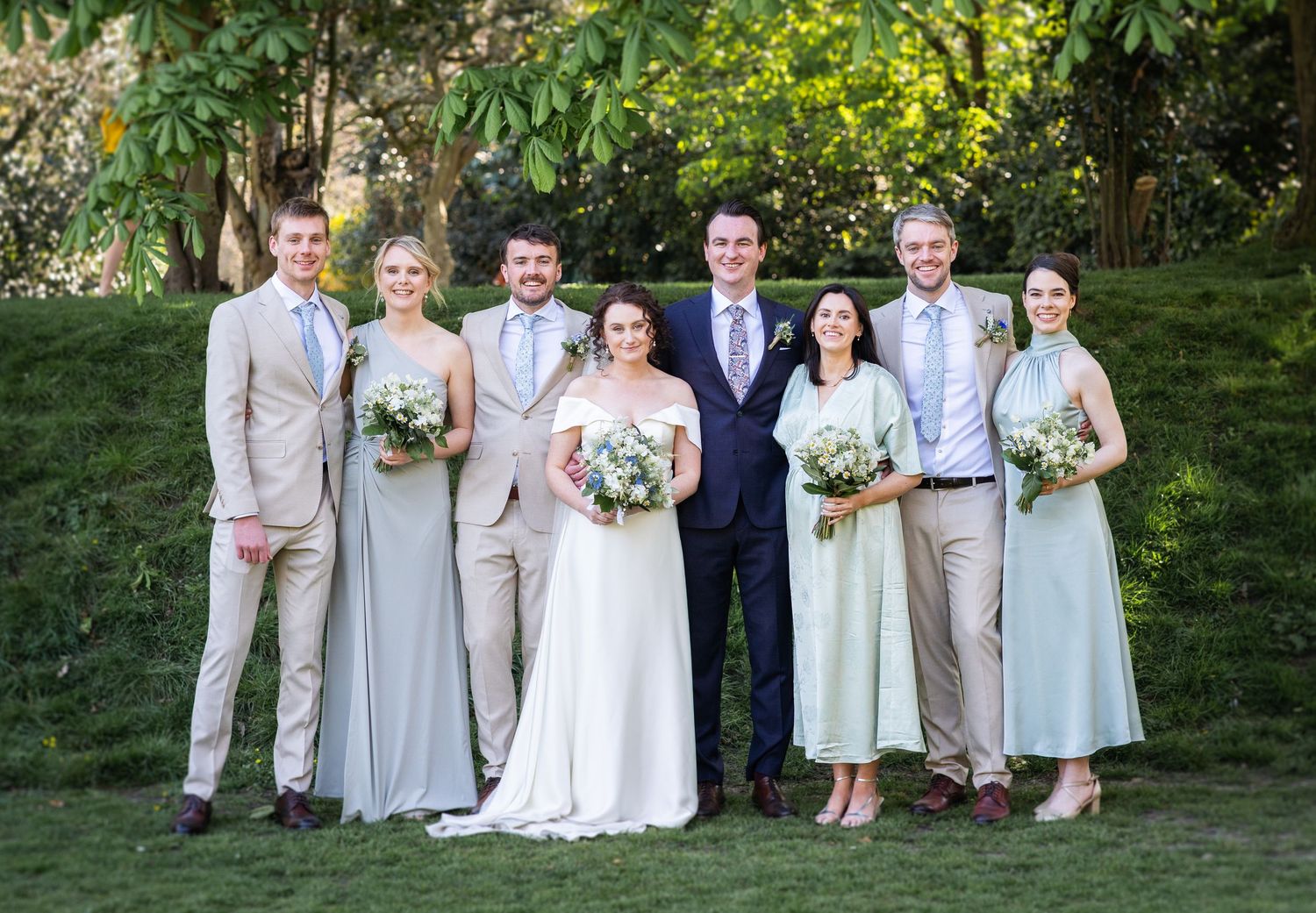 A wedding party in soft beige and blue attire poses outdoors against a lush green garden backdrop.