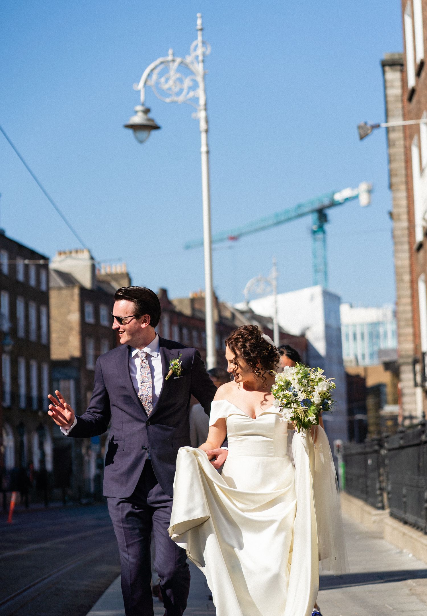 A couple in wedding attire walks down a city street with decorative lampposts and a construction crane in the background.