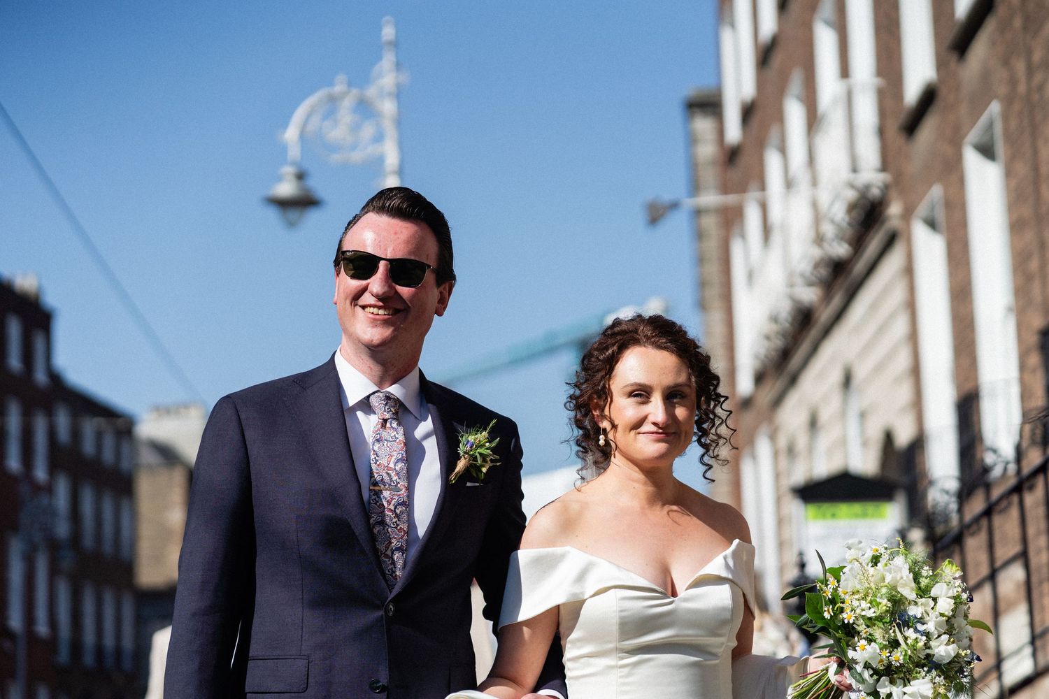 A couple walks down a sunny city street on their wedding day, the bride carrying a white bouquet.