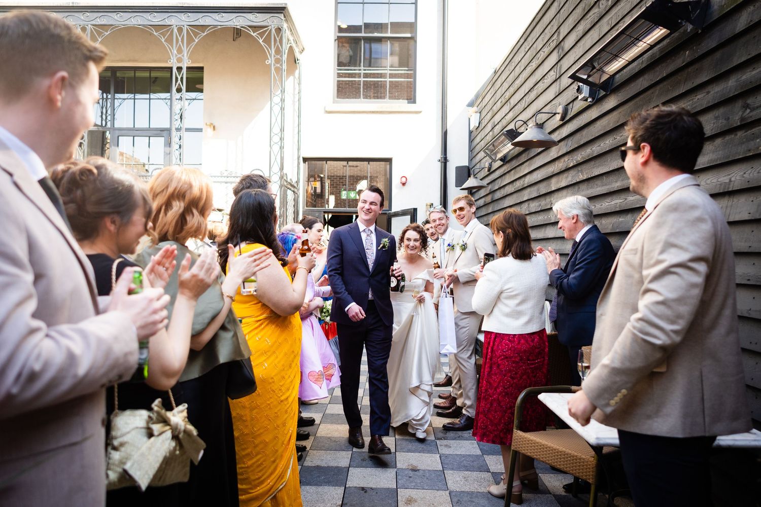 Wedding guests line both sides of a courtyard walkway to celebrate as newlyweds walk through the crowd.