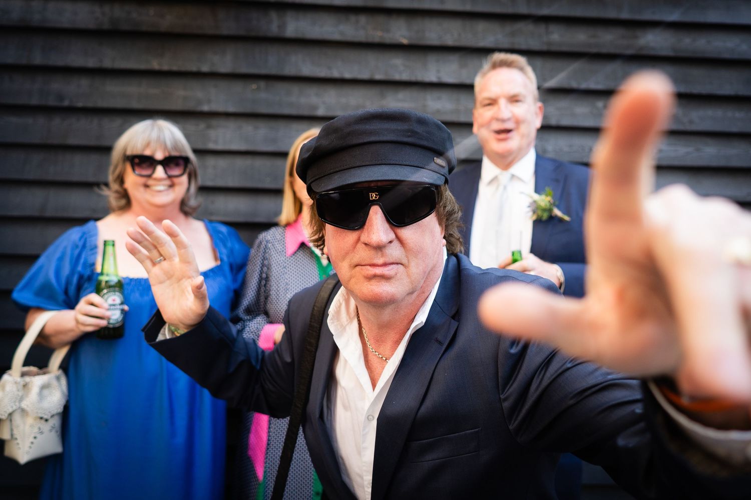 People celebrate at an outdoor event with one wearing sunglasses and a nautical cap making gestures at the camera.