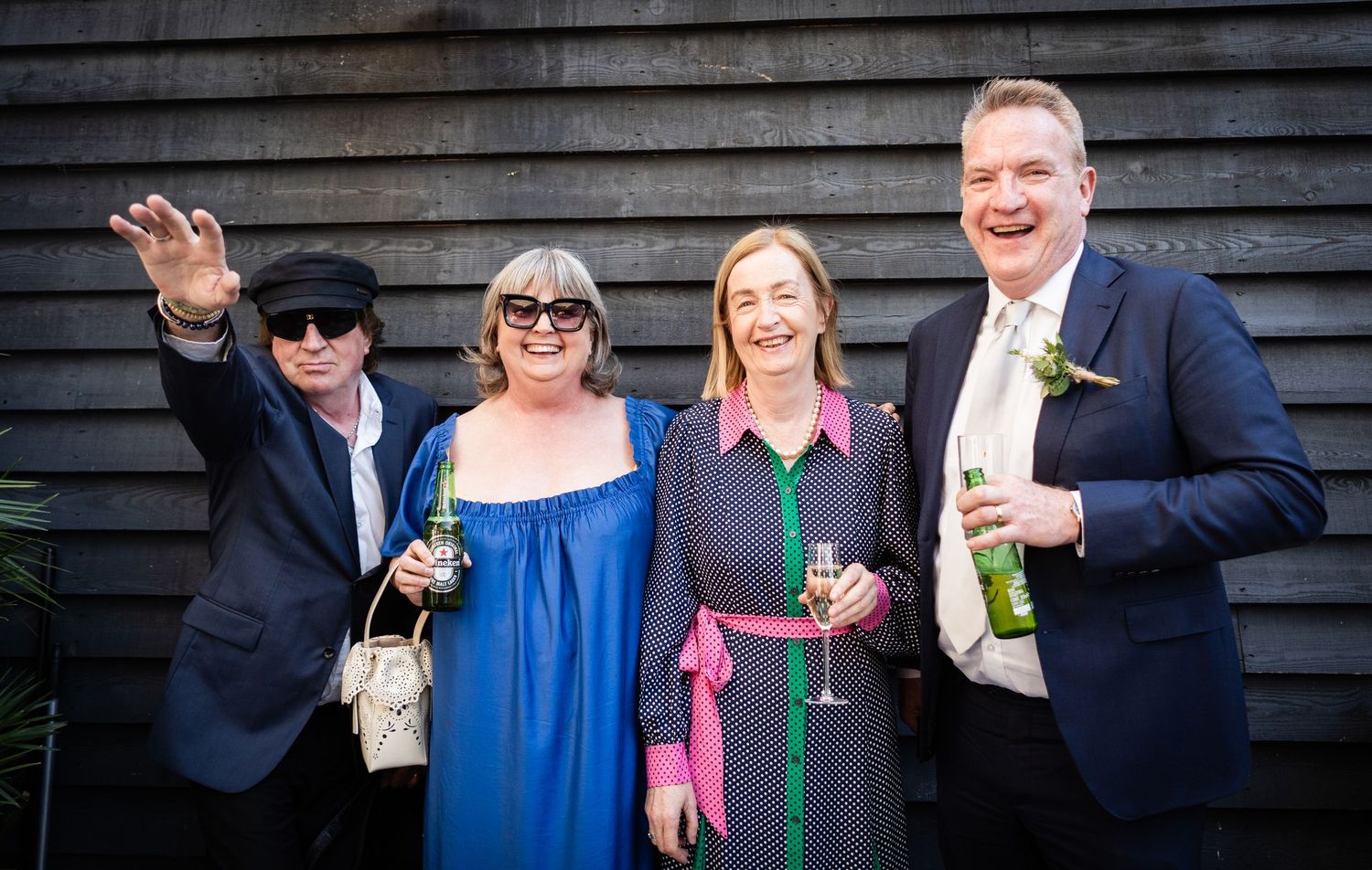Group of wedding guests smiling and posing together outdoors by a wooden wall while holding drinks.