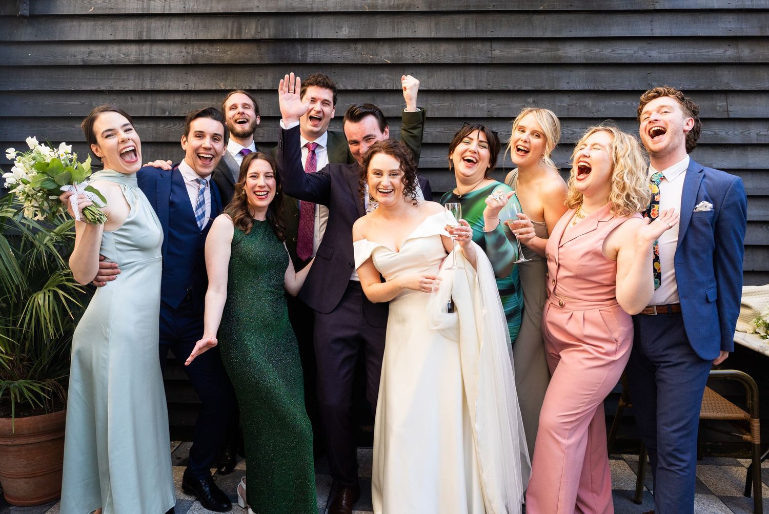 Wedding party in formal attire laughing together in front of wooden wall wearing colorful dresses and suits.