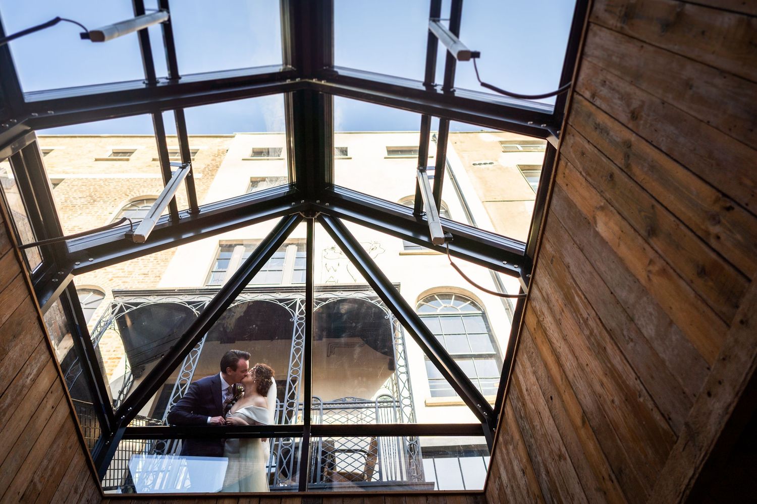 Looking up through a wedge-shaped glass skylight with wooden beams in an industrial-style building interior.