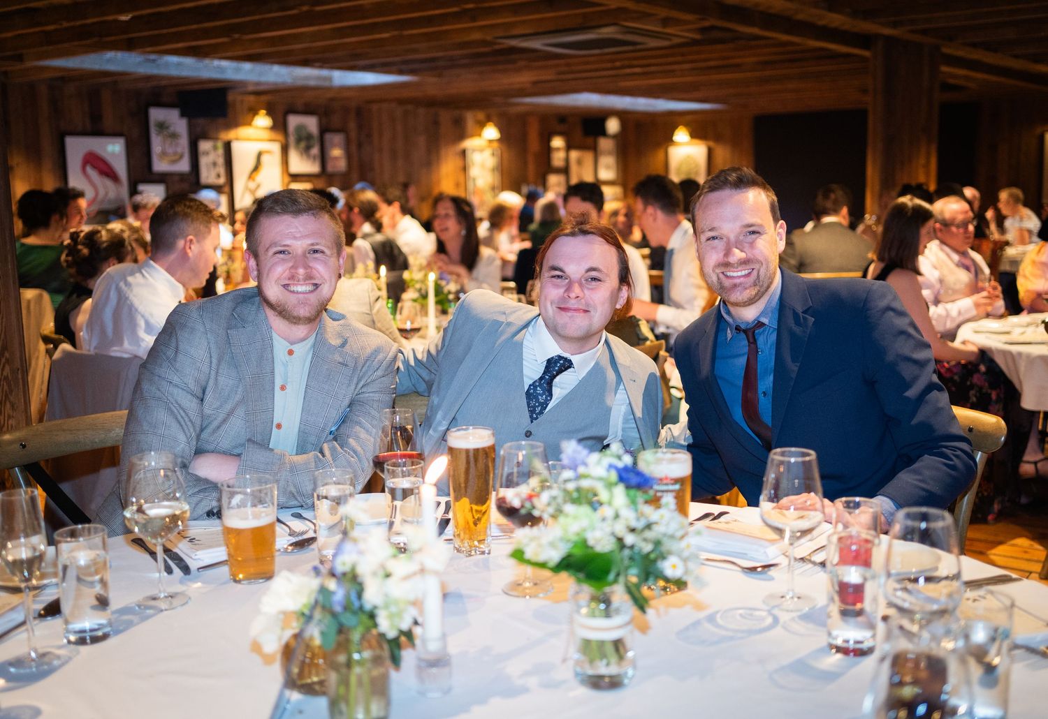 Guests enjoy a celebratory dinner with drinks at an elegant rustic venue with warm lighting and floral centerpieces.