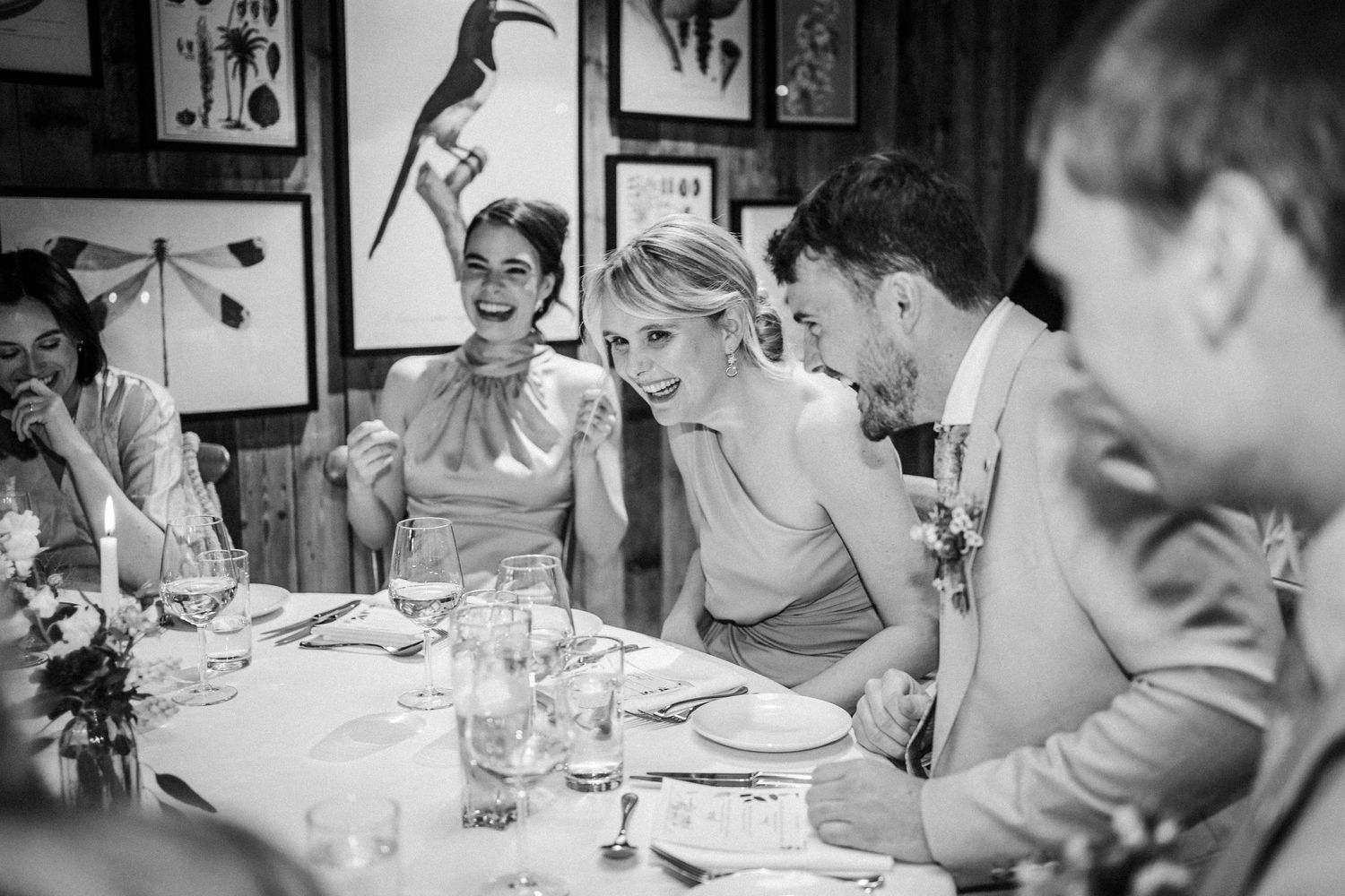 Black and white photo of wedding guests sharing laughter at an elegant dinner table with framed art on the walls.