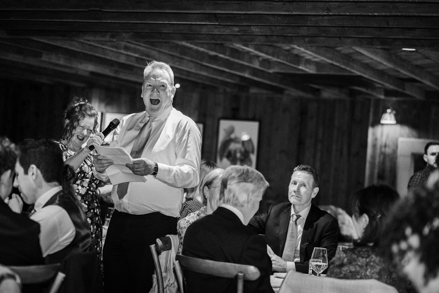 A black and white candid photograph of a wedding guest speaking during a reception dinner at a dimly lit indoor venue.