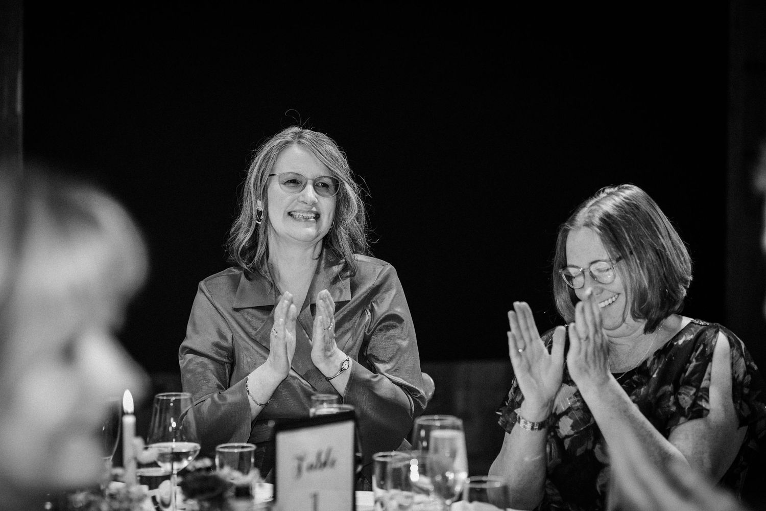 Black and white photo of people laughing together at a dinner table, showing genuine joy and connection during a social gathering.