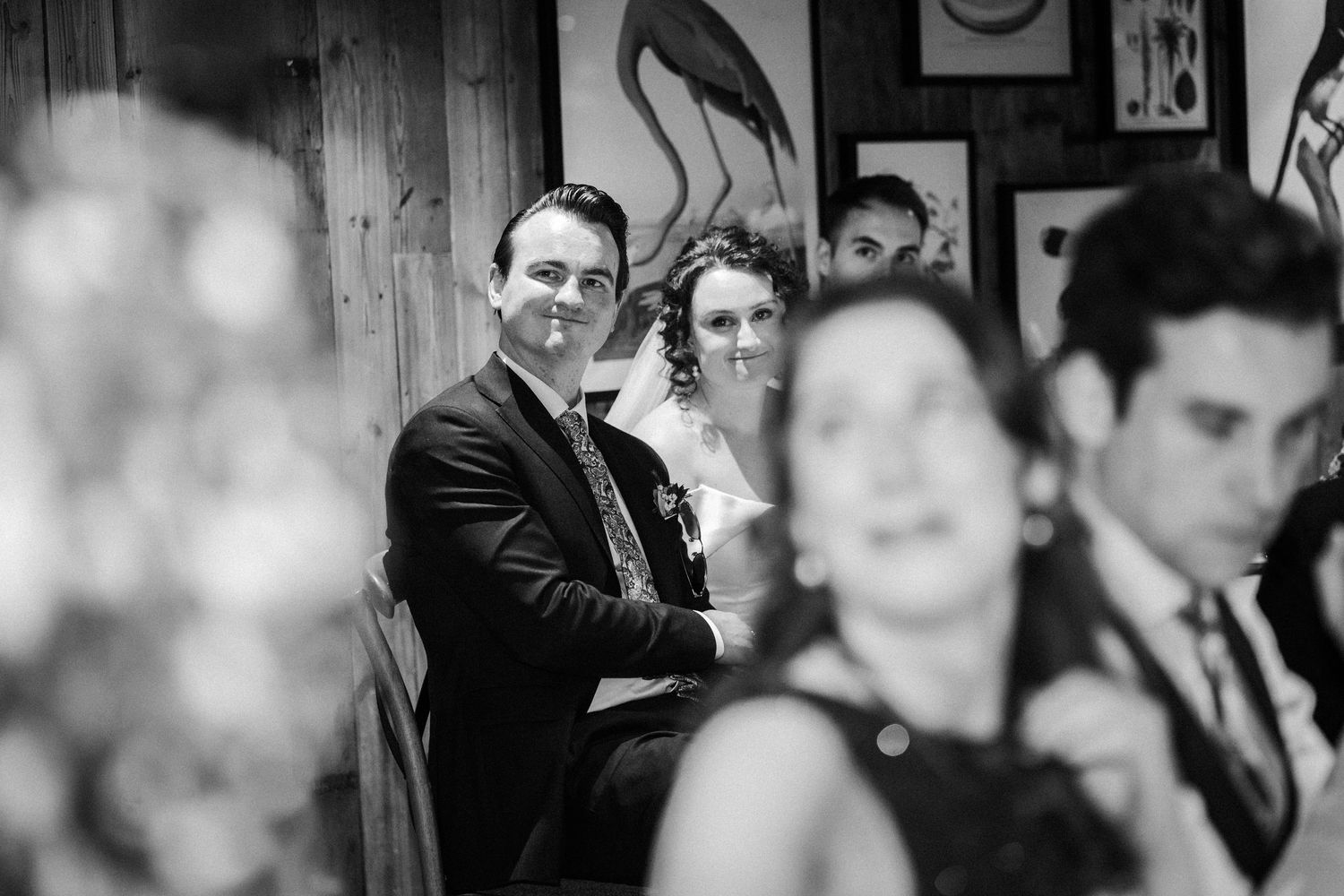 A black and white candid photograph of wedding guests at a reception with rustic wood paneling and framed artwork visible.