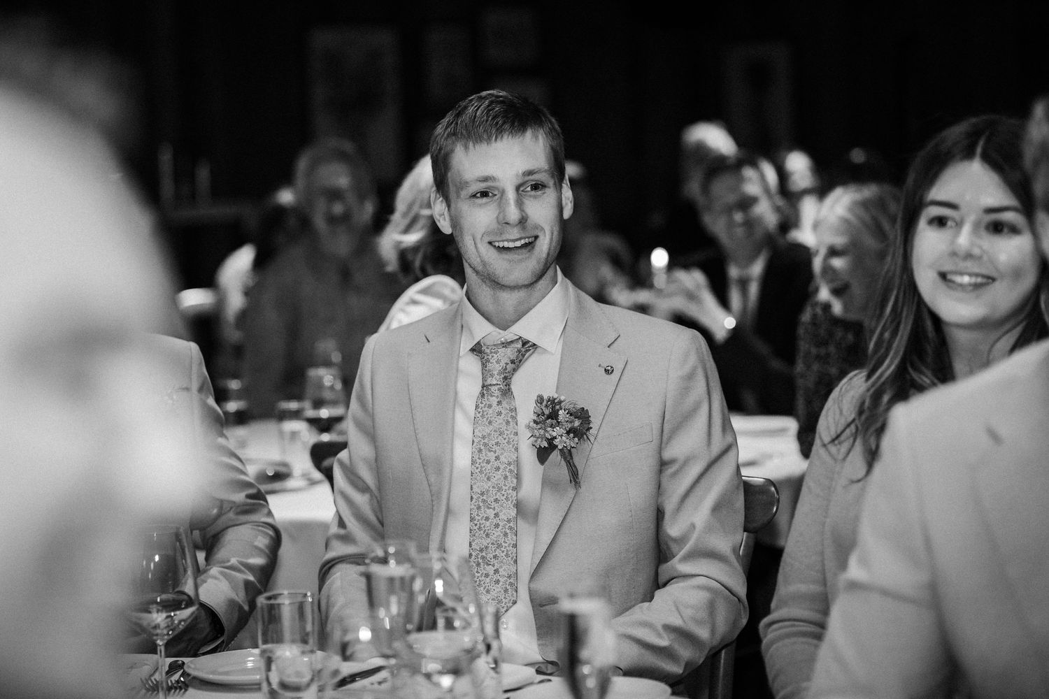 Black and white photo of guests smiling at formal event with table settings and decorations.