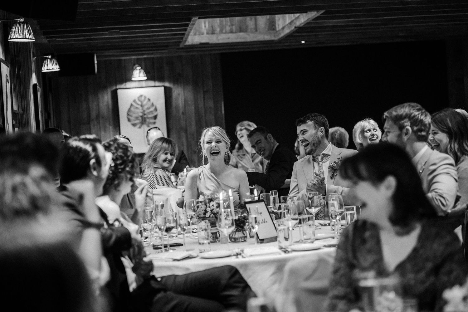 Wedding reception guests gather at a dinner table in a dimly lit restaurant, captured in black and white photography.