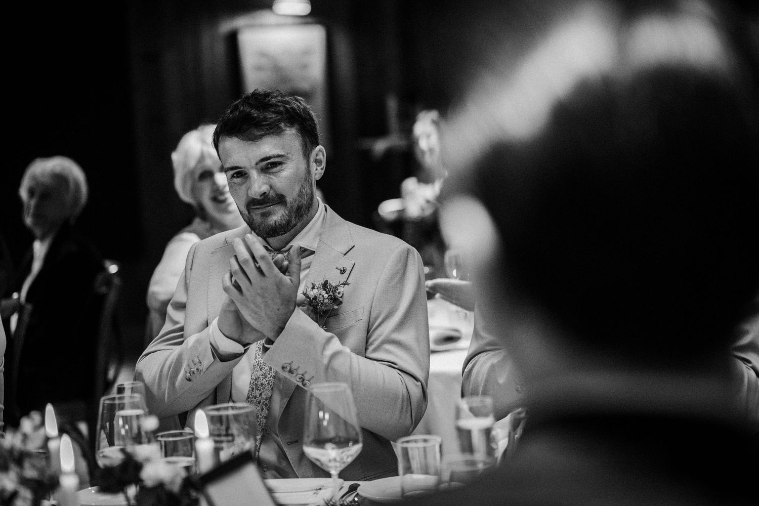 A wedding guest sits at a reception table, laughing during an emotional black and white moment.