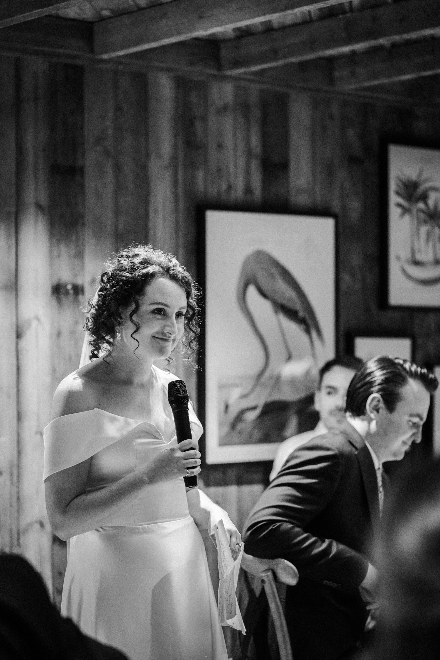 A black and white candid photograph at a wedding reception showing a speaker addressing guests beside artwork on wooden walls.