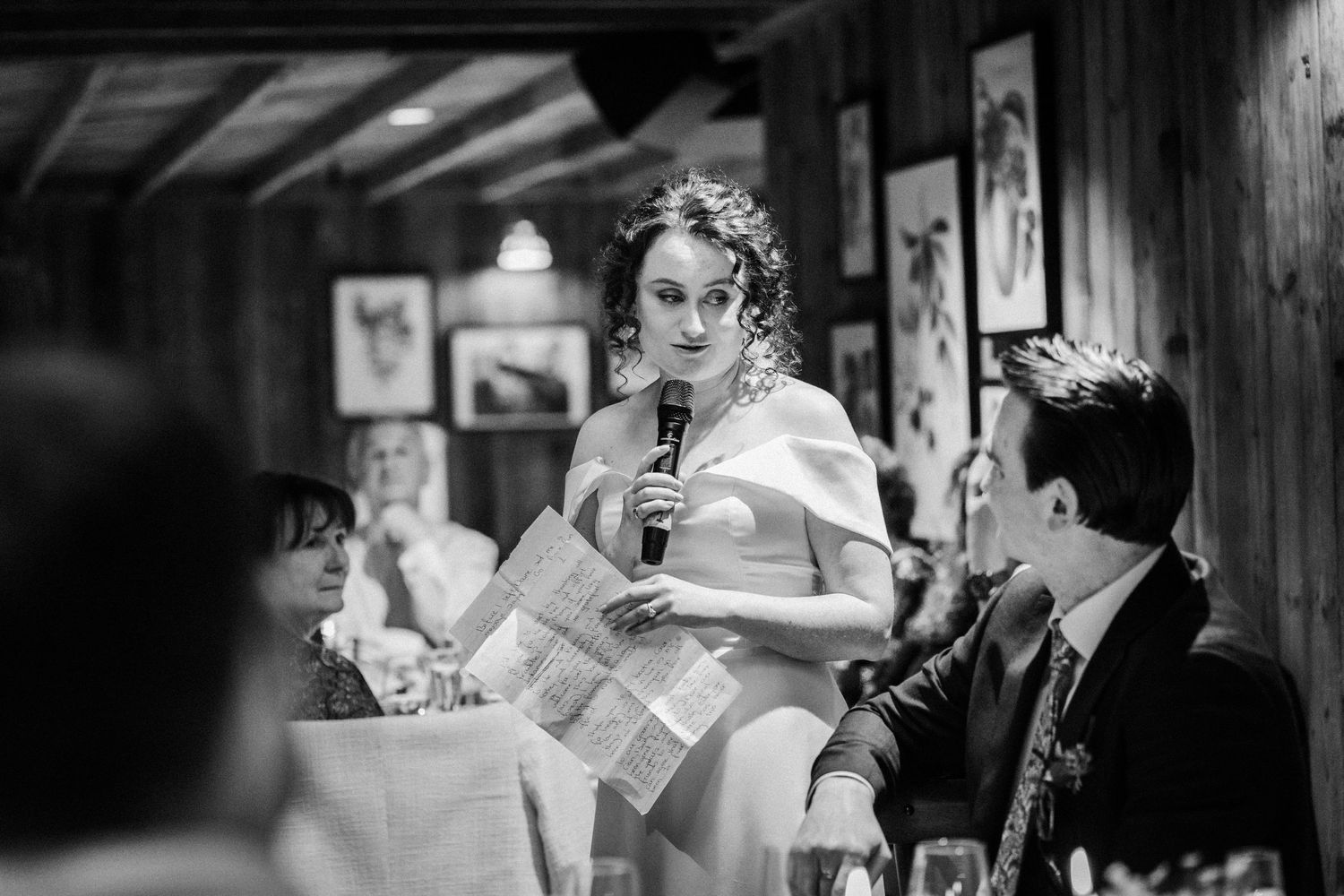A black and white photograph of someone giving a speech at a reception while holding papers and a microphone.