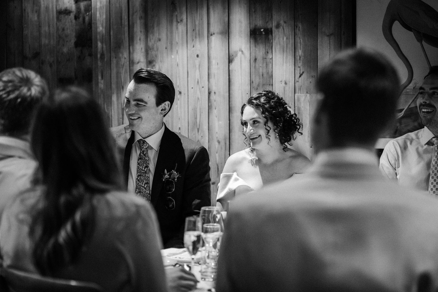 Black and white candid photo of wedding guests seated at a reception table enjoying conversation and laughter.