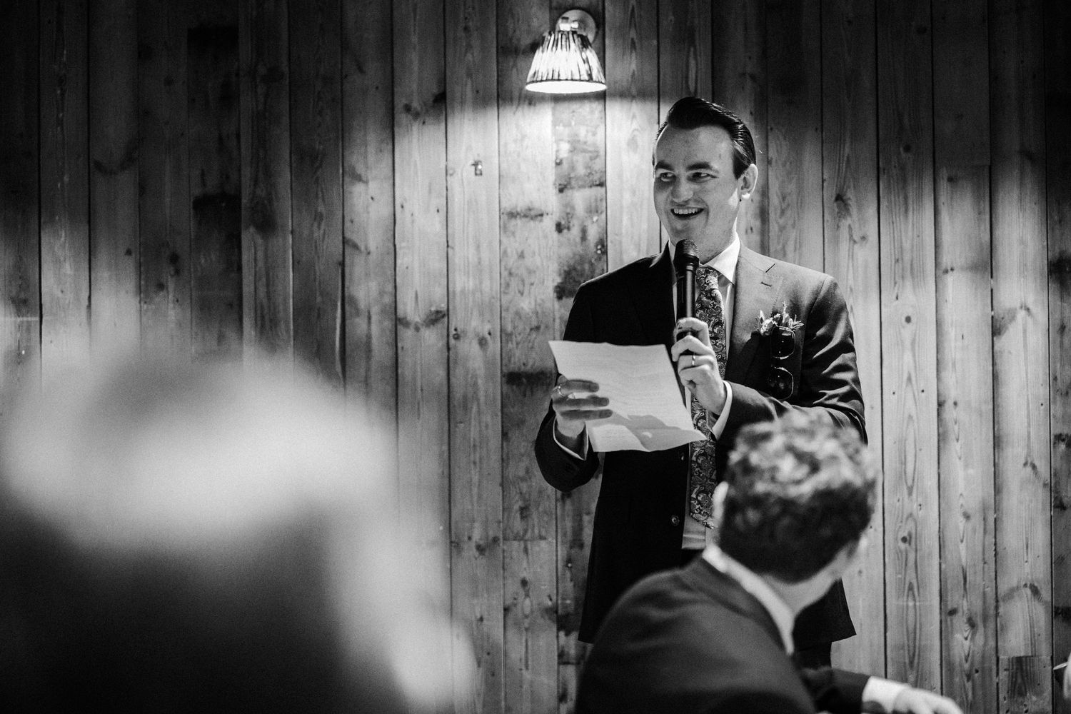 Black and white photo of a speaker reading from papers during a wedding ceremony with wooden wall background.