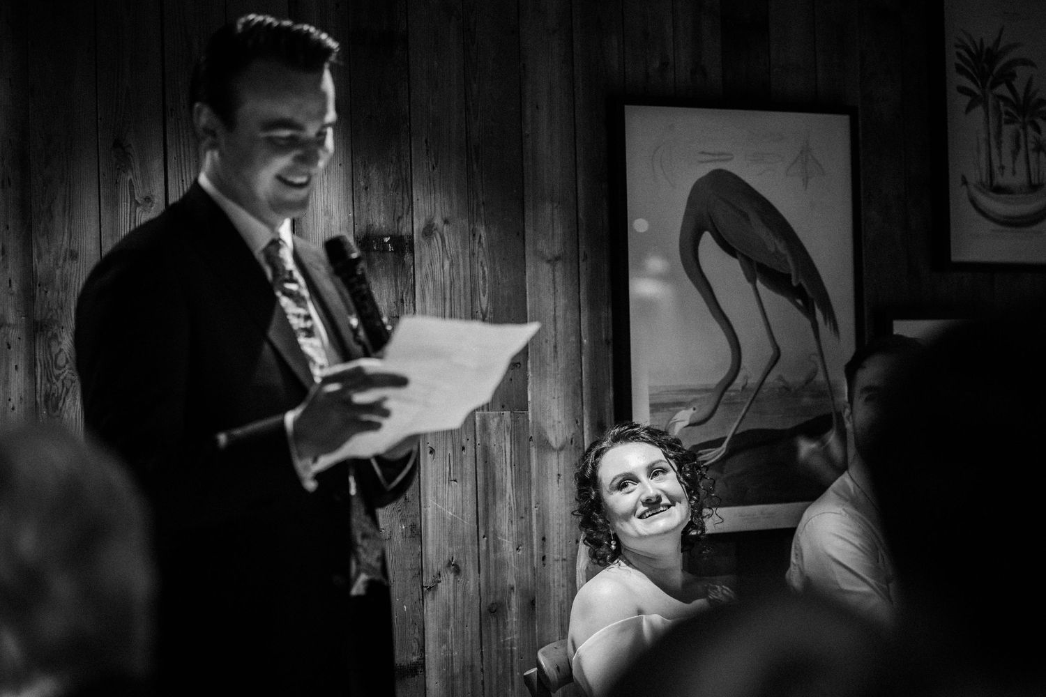 Black and white wedding reception scene with someone reading a speech while guests smile in a rustic wood-paneled room.