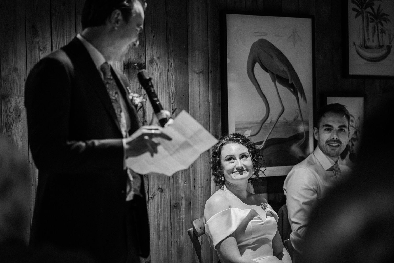A black and white wedding ceremony moment with guests seated in front of a wall decorated with a vintage bird illustration.
