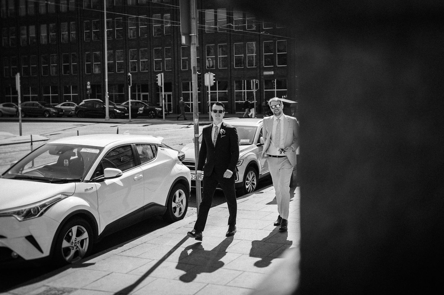 A monochrome street scene with people walking past parked cars outside brick buildings.