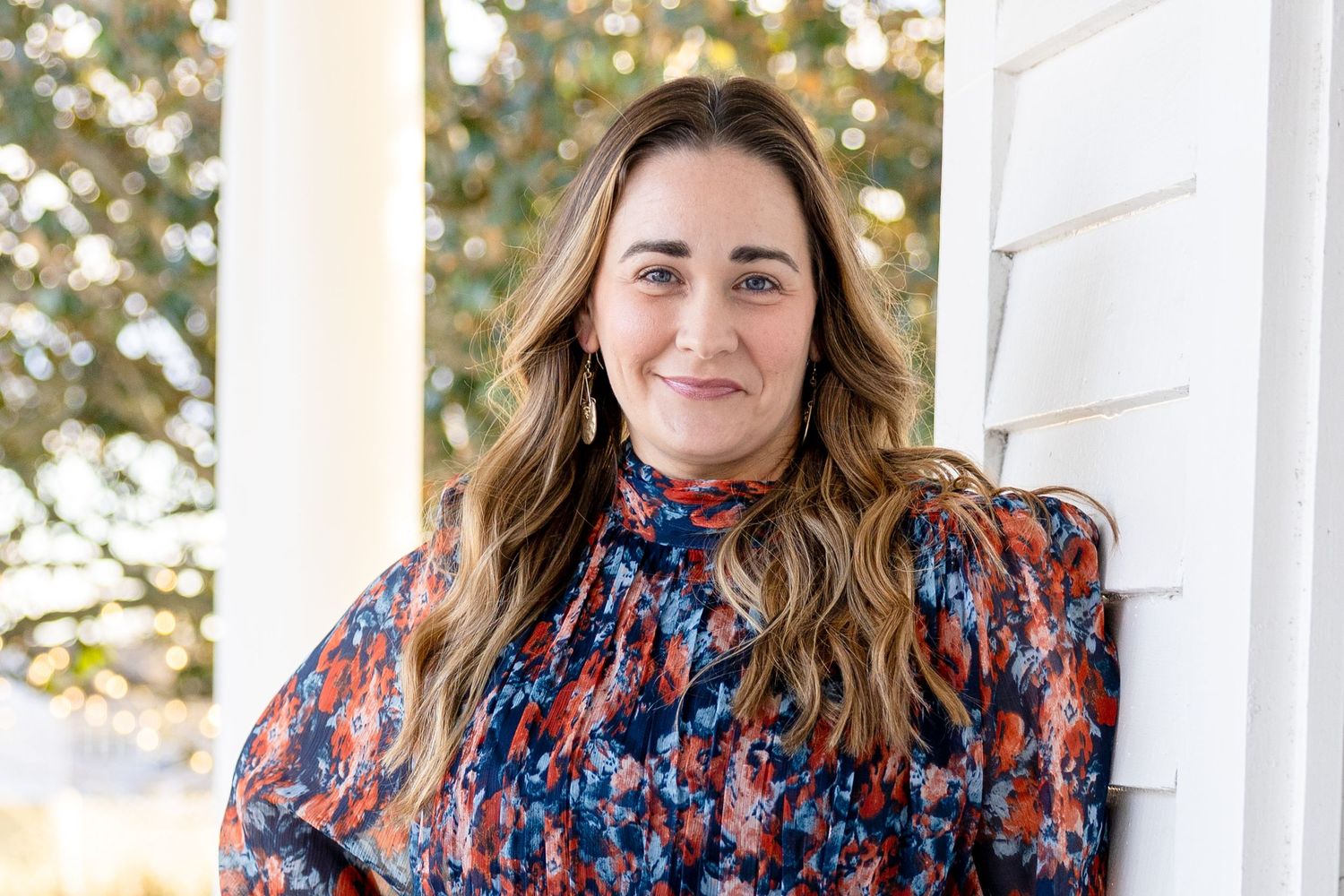 Someone wearing a colorful patterned blouse sits outdoors against a white wall with blurred foliage in the background.