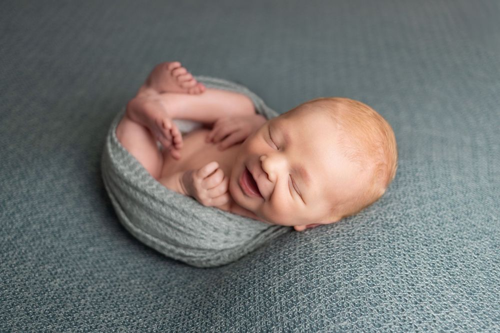 A peaceful newborn baby swaddled in gray fabric sleeps curled up on a textured gray backdrop.