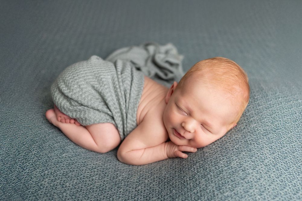 Newborn baby sleeping peacefully on gray fabric while wearing gray wrapping.