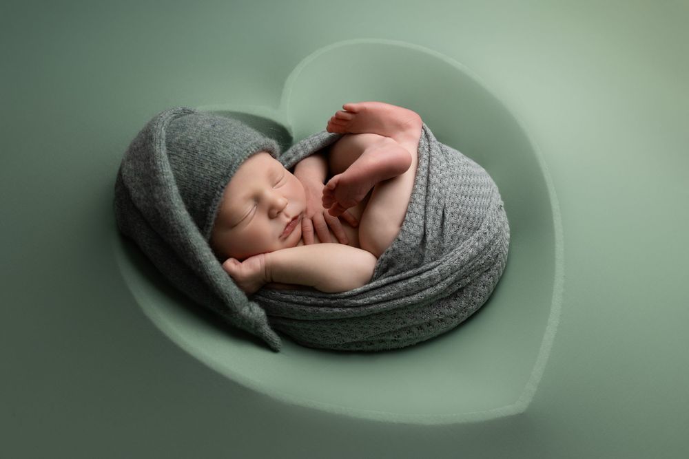 A sleeping newborn baby peacefully curled up in gray knit blanket on sage green heart-shaped backdrop.