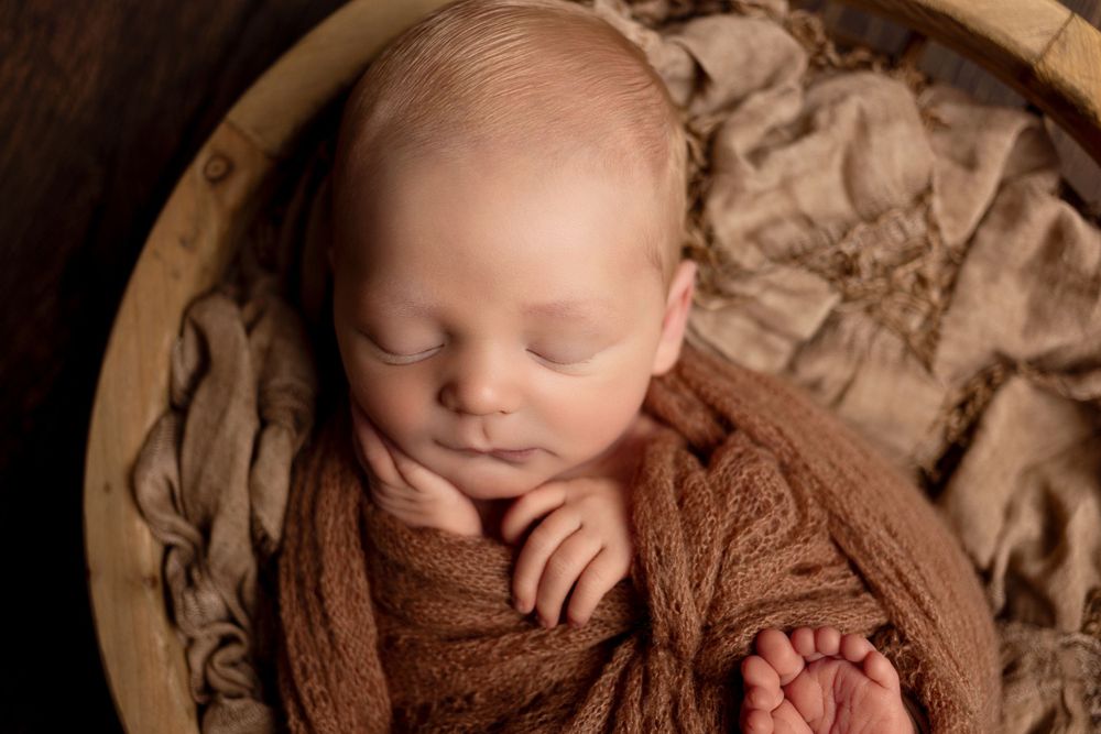 A sleeping newborn baby wrapped in brown fabric lies peacefully in a wooden bowl.