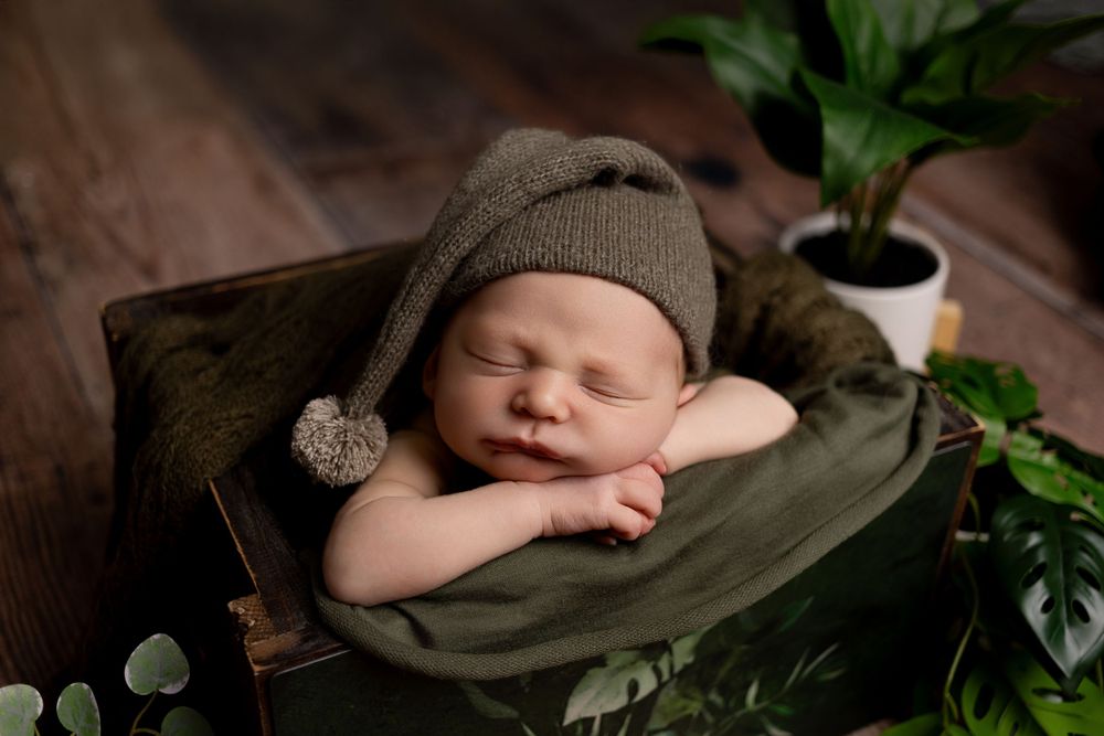 Peaceful newborn sleeping on olive green blanket surrounded by green plants in studio photography setup.