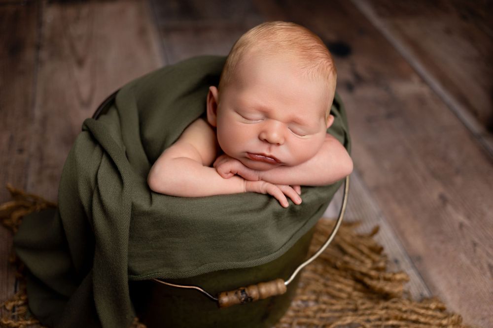 Sleeping newborn baby wrapped in olive green fabric resting peacefully on rustic wooden floor.