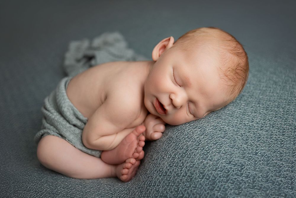 A sleeping newborn baby curled up in fetal position on gray fabric with soft natural lighting.