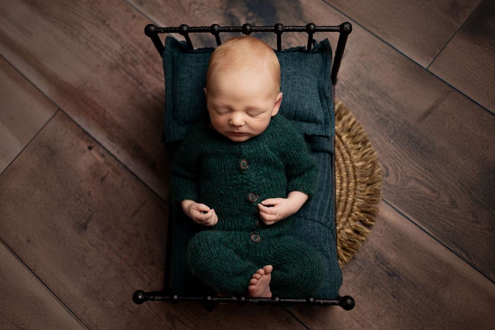 Sleeping newborn in dark green wrap rests peacefully on black hammock against wooden floor backdrop.