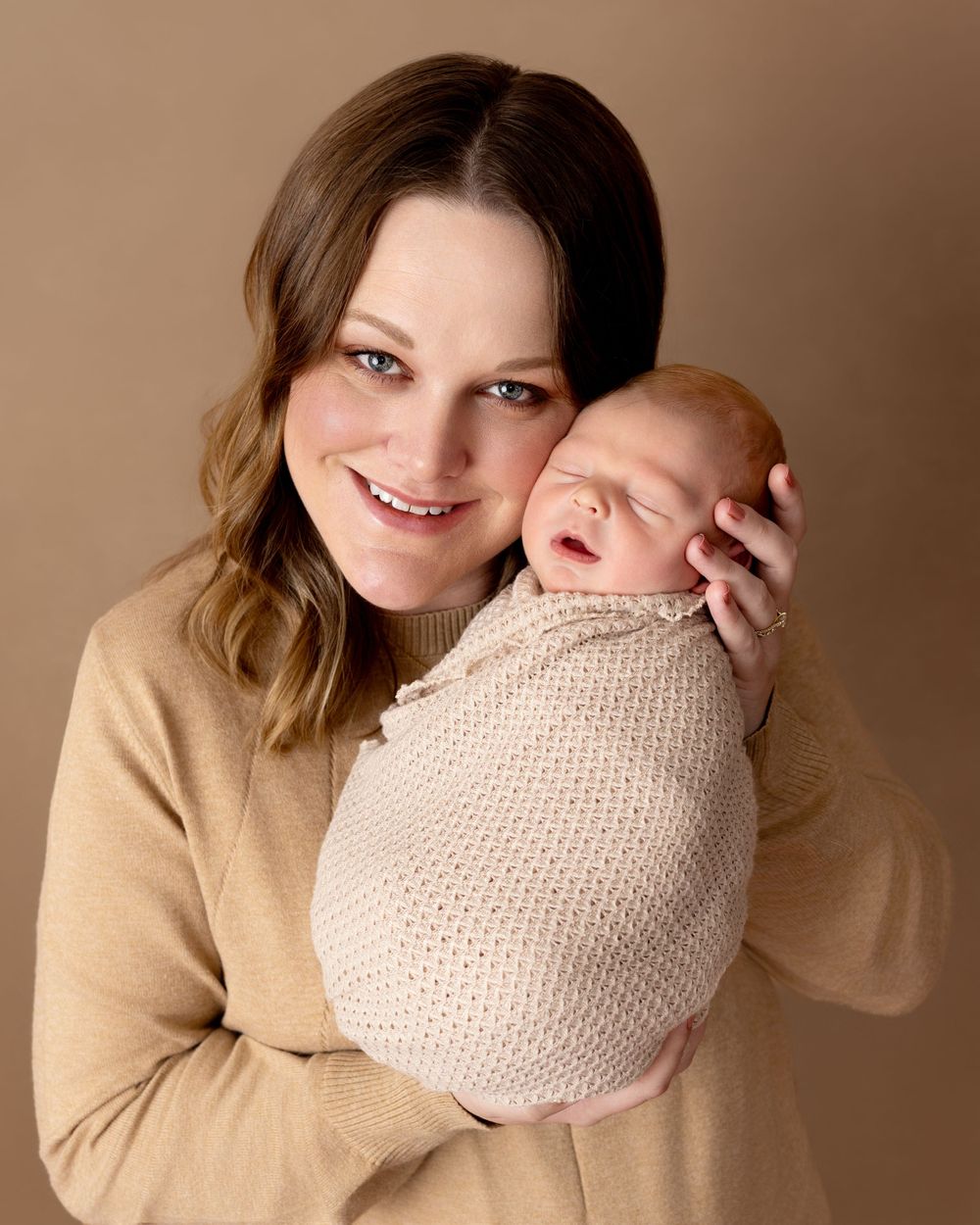 A person in a tan sweater holds a smiling newborn baby wrapped in a beige knit blanket in a warm studio portrait.