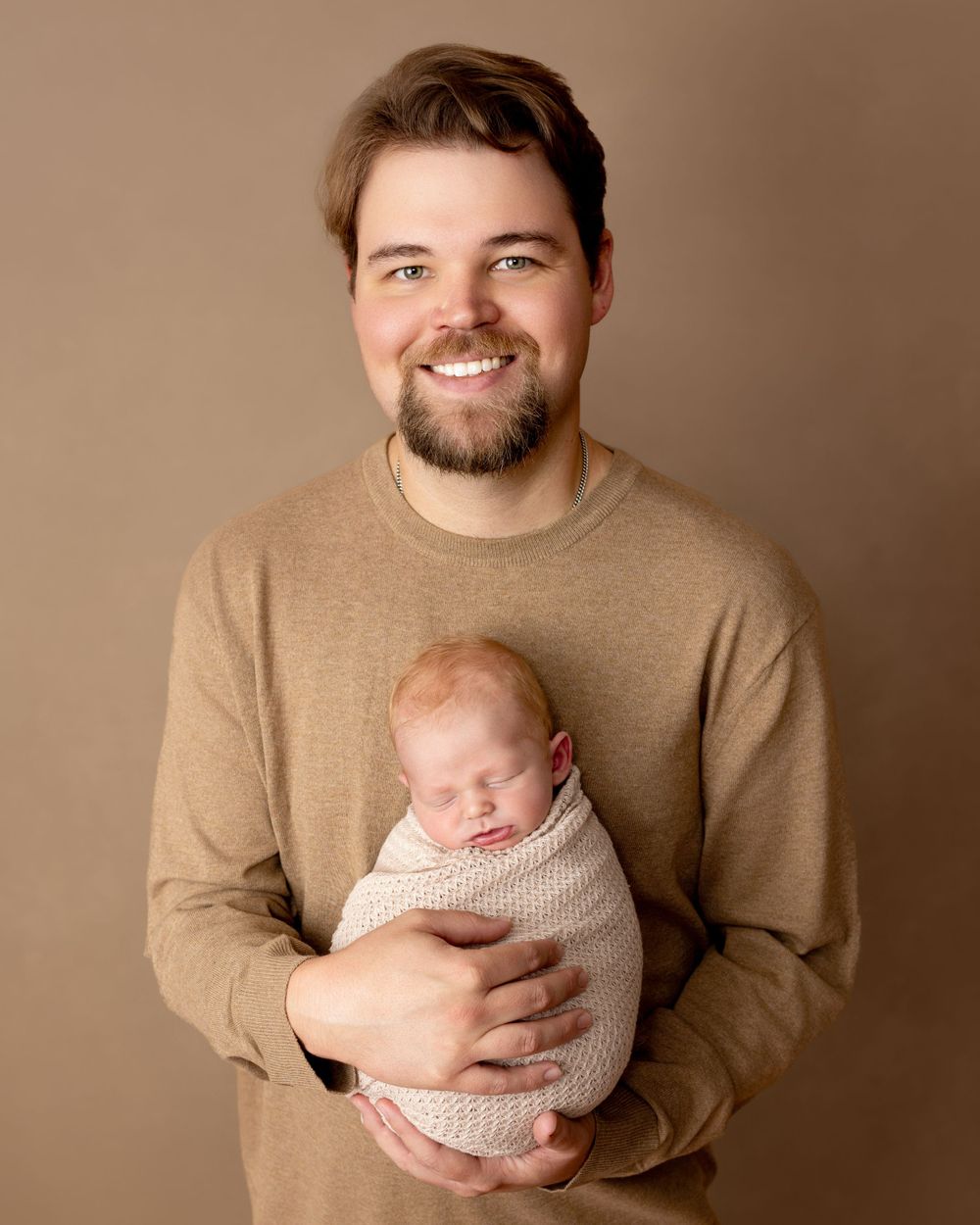 A person in a beige sweater gently holds a sleeping newborn baby wrapped in a soft cream-colored blanket.