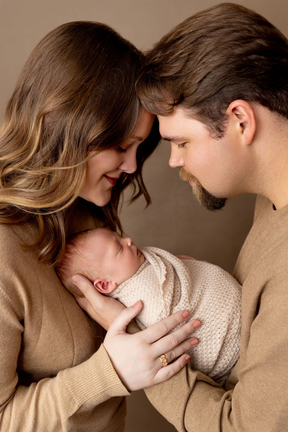 Loving couple in tan sweaters tenderly holding their newborn baby wrapped in knit blanket against warm brown background.