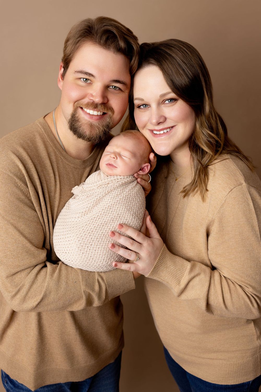 A couple in matching tan sweaters holds their sleeping newborn baby wrapped in a knit blanket against a neutral backdrop.