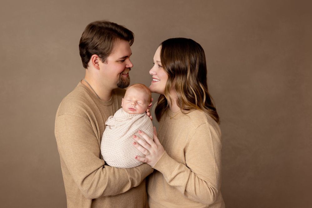 Family wearing matching beige sweaters pose together with newborn baby against neutral brown studio backdrop.