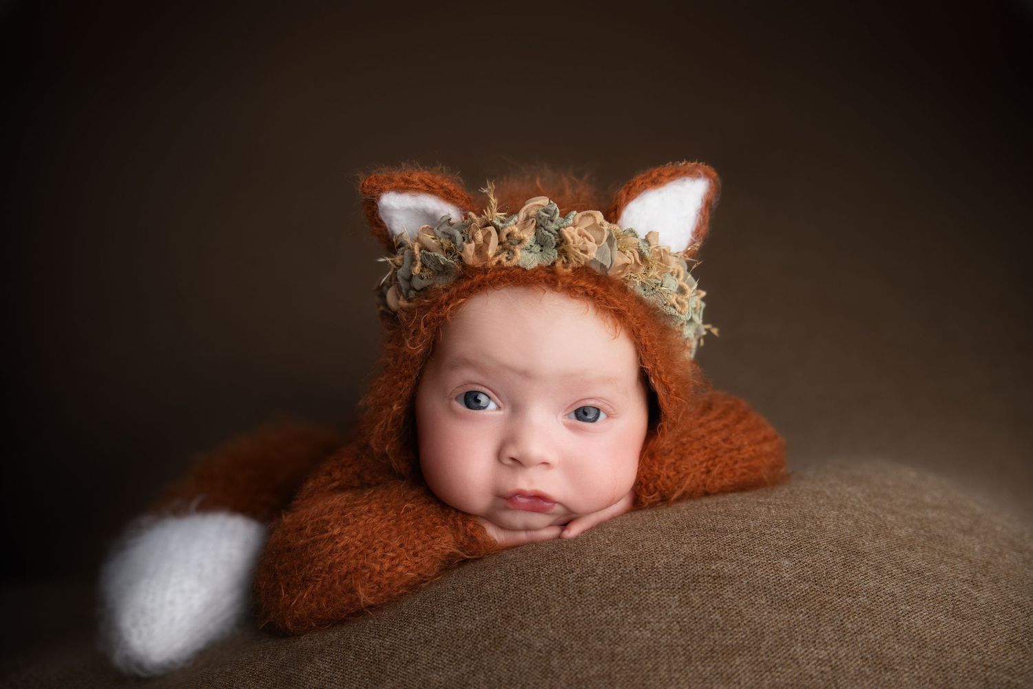 Baby wearing a brown fox costume with floral crown peeks out from behind a soft surface.