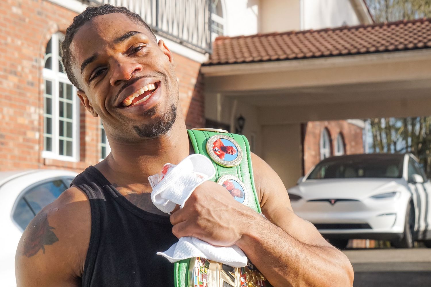 A person in a black tank top smiles while holding a pack of ginger ale outside a house with Spanish tile roof.